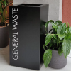 black office recycling bin with white lettering General Waste. by pot plant