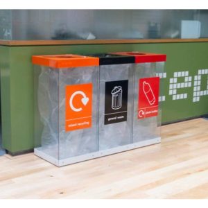 3 transparent office recycling bins by a reception. Orange Mixed Recycling label and top, Black General Waste and top and red Plastic Bottles label and top