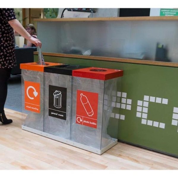 3 transparent office recycling bins in front of a reception with orange top and Mixed Recycling label, Black Top and General Waste label and red top and Plastic Bottles label